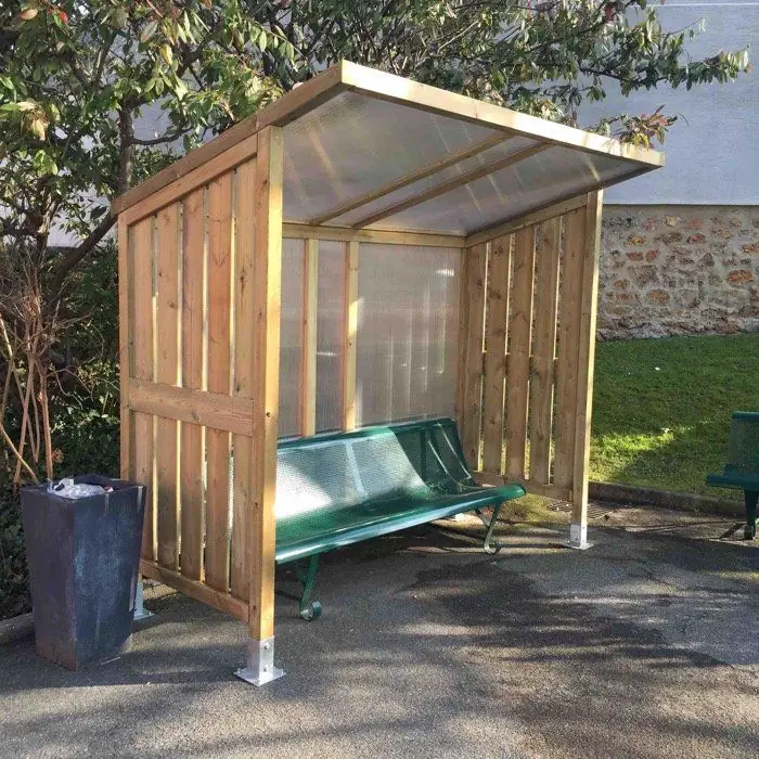 Abri en bois couvert de polycarbonate abritant un banc, dans un parc