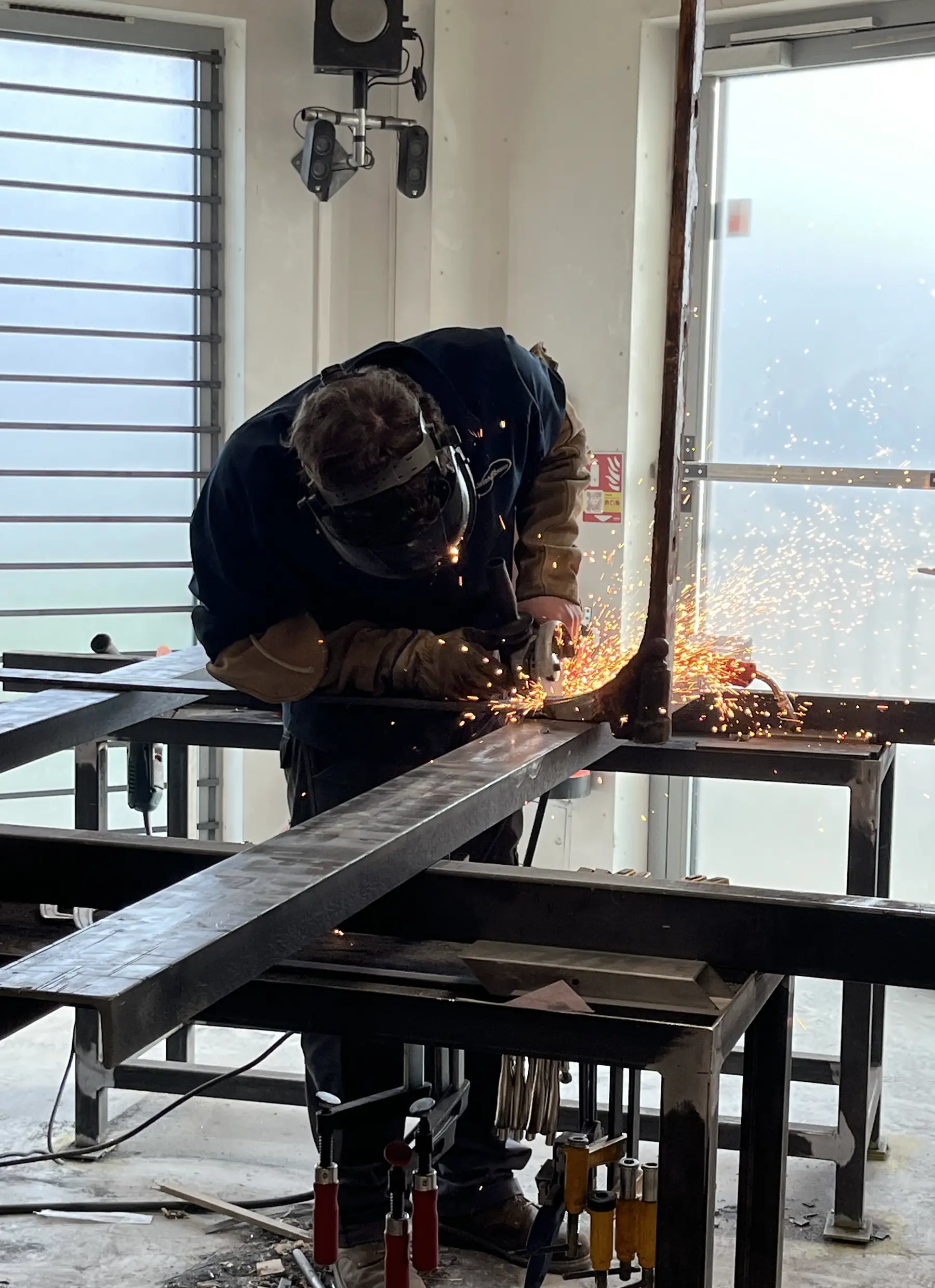 Compagnon meulant un profilé en acier sur un établi de l’atelier, gerbe d’étincelles